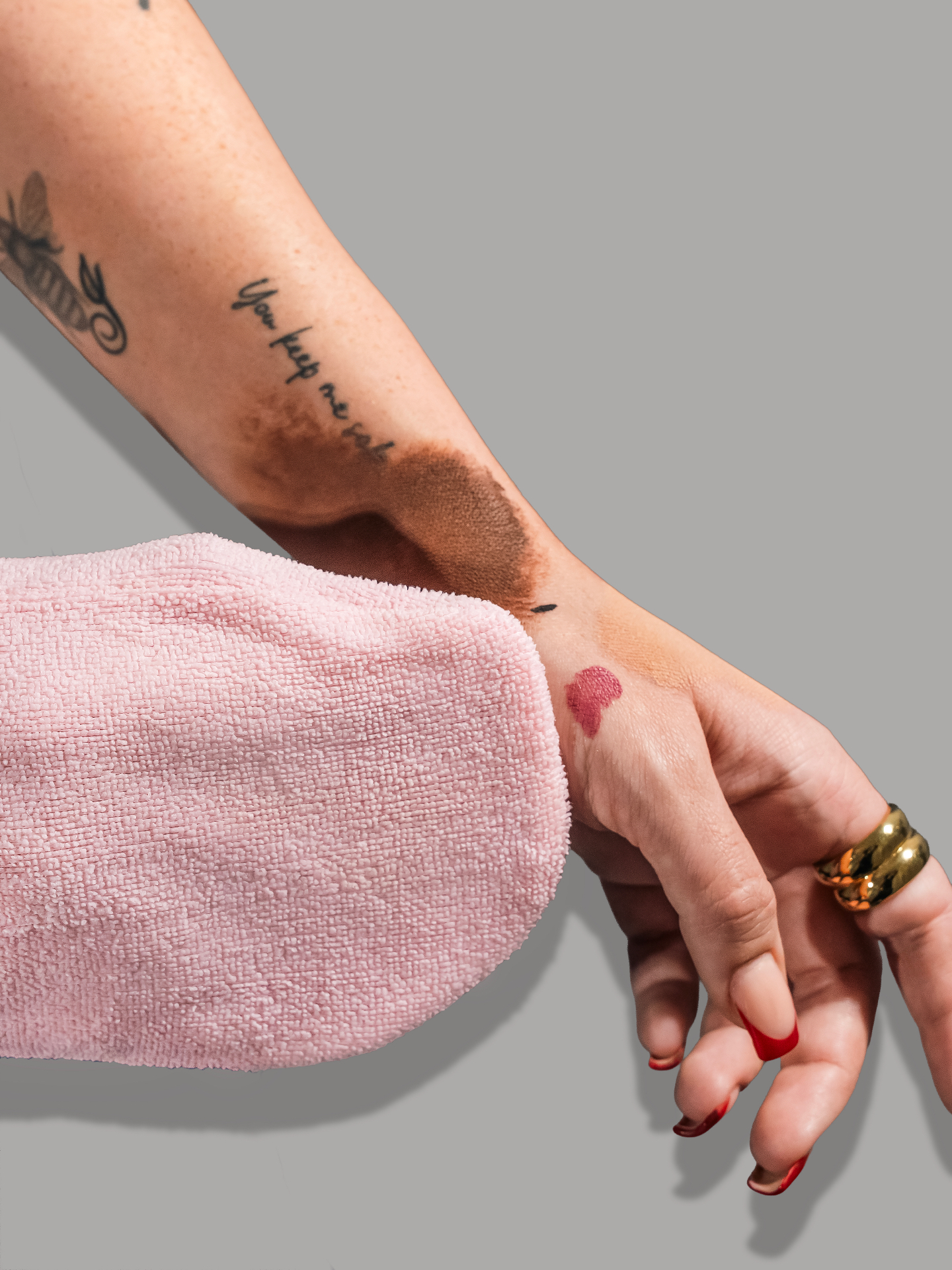 A pink Smitten mitt wiping foundation and lipstick swatches off a woman's forearm, showing how the cloth lifts pigment away from skin