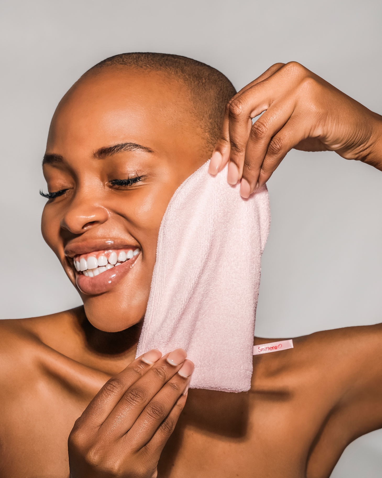 A smiling woman with a closely shaved head holding a soft pink Smitten cloth up to her cheek with both hands
